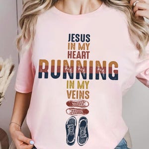 May include: Pink t-shirt with the text "Jesus in my heart running in my veins" in a vintage style font. The text is in shades of brown, orange, and blue. There are two pairs of running shoes printed below the text.