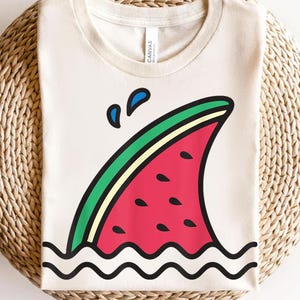Puede incluir: Una camiseta blanca con un gráfico de una sandía con forma de aleta de tiburón, con gotas de agua encima. La sandía es roja con semillas negras y una cáscara verde. El agua se representa como olas azules.