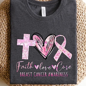May include: A gray t-shirt with a pink ribbon, heart, and cross design. The design is made up of pink ribbons with floral patterns. The text "Faith love cure" and "Breast Cancer Awareness" is printed below the design.