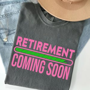 May include: A gray t-shirt with the words "RETIREMENT COMING SOON" in pink, with a green loading bar graphic. The shirt is laid on top of blue jeans and a tan hat.
