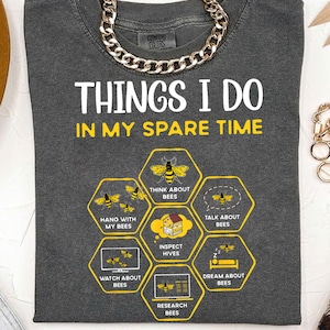 May include: A grey t-shirt with the text "Things I do in my spare time" and a honeycomb graphic with bee-related activities, including "Think about bees", "Hang with my bees", "Talk about bees", "Inspect hives", "Watch about bees", "Research bees", and "Dream about bees".
