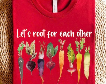 Könnte beinhalten: Rotes T-Shirt mit dem Text "Let's root for each other" in weiß gedruckt. Das Shirt zeigt eine Reihe bunter Gemüse, darunter Karotten, Radieschen und Rote Bete.