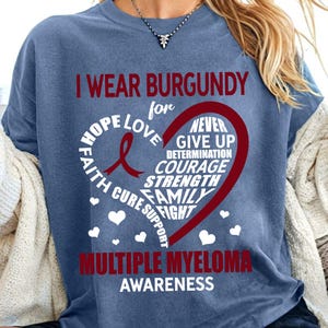 May include: A blue t-shirt with the words "I wear burgundy for" and a red heart with the words "Hope, Love, Faith, Cure, Support, Never Give Up, Determination, Courage, Strength, Family, Fight" inside. The words "Multiple Myeloma Awareness" are printed below the heart.