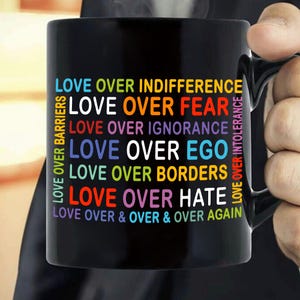 May include: Black ceramic mug with the words "LOVE OVER" followed by various terms like "INDIFFERENCE," "FEAR," and "HATE" in colorful text. The mug also has text on the side that says "LOVE OVER BARRIERS" and "LOVE OVER INTOLERANCE."