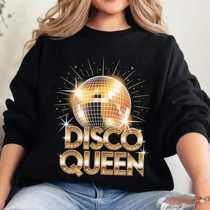 May include: Black sweatshirt featuring a gold disco ball graphic with starbursts and the text "DISCO QUEEN" in gold block letters. The design has a retro, vintage aesthetic. The sweatshirt is a casual, comfortable style.