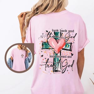 Puede incluir: Camiseta rosa claro con un dise&ntilde;o de cruz, un coraz&oacute;n rosa y las palabras "look back and thank God, look forward and trust God". El dise&ntilde;o incluye detalles verdes, negros y amarillos.