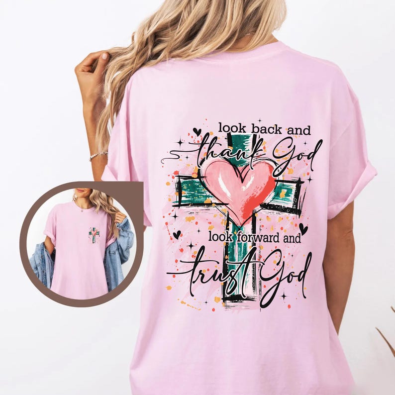 Puede incluir: Camiseta rosa claro con un dise&ntilde;o de cruz, un coraz&oacute;n rosa y las palabras "look back and thank God, look forward and trust God". El dise&ntilde;o incluye detalles verdes, negros y amarillos.