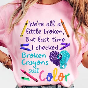 May include: Pink t-shirt with a colorful design featuring crayons, a heart, and the text "We're all a little broken, But last time I checked Broken Crayons still Color".