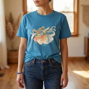 May include: A light blue t-shirt with a watercolour illustration of a whirling dervish. The design depicts flowing robes in peach, yellow, and blue. The shirt is made of a soft material and has a classic crew neck.