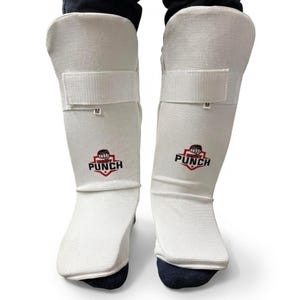 May include: Pair of white shin guards with a hook-and-loop strap and the word "M" printed on the strap. The "BODY PUNCH" logo is printed in red and black on the front of each guard. The guards are worn over dark socks.