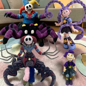 Puede incluir: Una colección de muñecos de peluche hechos a mano con diseños únicos. Los muñecos incluyen una figura tipo bufón con cuerpo de araña, una niña con trenzas moradas y otros personajes caprichosos. Los muñecos son coloridos y detallados, con varias texturas.