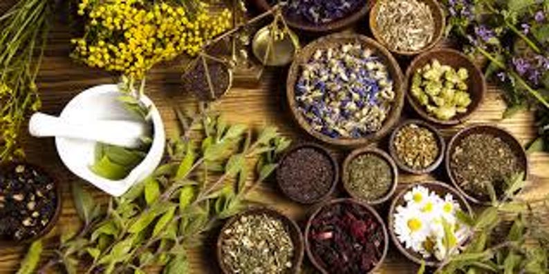 May include: A collection of dried herbs and flowers in small bowls, arranged on a wooden surface. The herbs and flowers are a variety of colors, including green, brown, yellow, and purple. There is also a white mortar and pestle in the image.