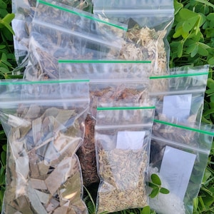 May include: Seven small clear plastic bags filled with various dried herbs and spices. The bags are laid out on a bed of green grass.