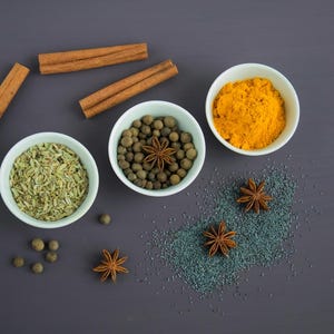 May include: A flat lay of various spices in small white bowls on a dark gray surface. The spices include fennel seeds, allspice berries, turmeric powder, and star anise.