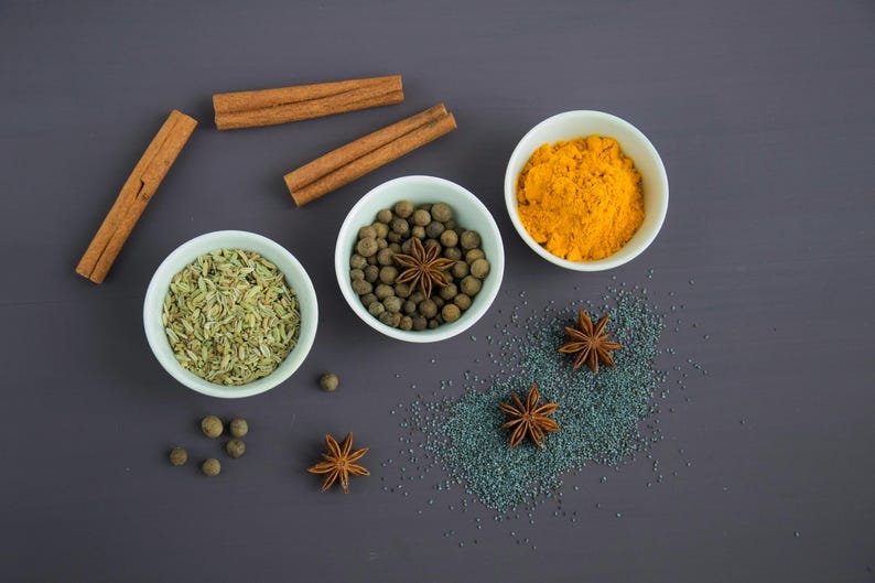May include: A flat lay of various spices in small white bowls on a dark gray surface. The spices include fennel seeds, allspice berries, turmeric powder, and star anise.