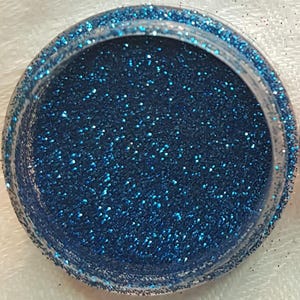 May include: A small, clear container filled with blue glitter. The glitter is fine and sparkly.