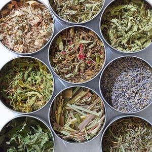 May include: Eight round metal containers filled with different dried herbs and flowers. The containers are arranged in a circle on a white surface. The herbs and flowers include lavender, lemongrass, mint, and other varieties.