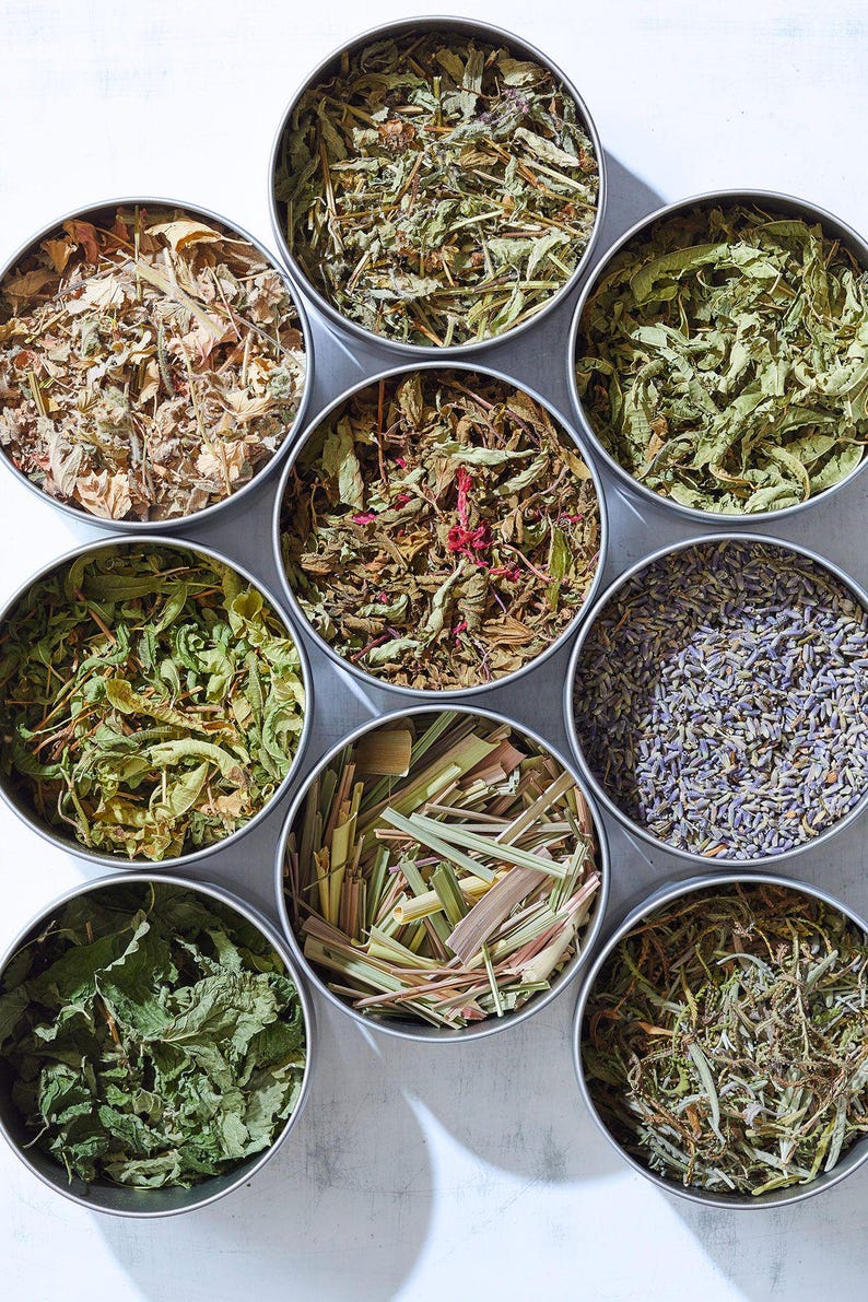 May include: Eight round metal containers filled with different dried herbs and flowers. The containers are arranged in a circle on a white surface. The herbs and flowers include lavender, lemongrass, mint, and other varieties.
