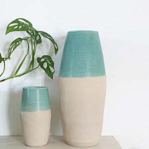 Può includere: Due vasi in ceramica dalla forma cilindrica affusolata. Ogni vaso presenta una smaltatura liscia e lucida color verde acqua nella parte superiore e una finitura beige opaca e testurizzata nella parte inferiore, creando un aspetto bicolore.
