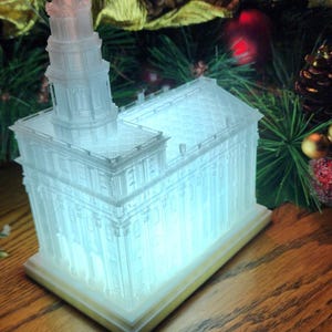 Op de afbeelding: Een wit, 3D-geprint model van een kerkgebouw met een licht erin, waardoor de structuur wordt verlicht. Het model staat op een houten oppervlak.