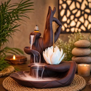 May include: A brown ceramic incense burner shaped like a hand, with a white lotus flower and a lit incense stick. The burner has a waterfall effect with smoke flowing down. A small candle and decorative stones are in the background.