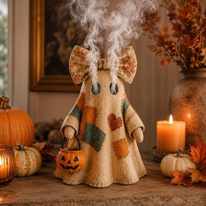 May include: A beige fabric ghost figurine featuring a large floral print bow on its head, patchwork accents in orange and green, and two dark eye holes emitting white smoke. The ghost holds a small orange plastic jack-o'-lantern bucket.