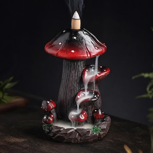 May include: A mushroom-shaped incense burner with a red and black cap, white polka dots, and a brown stem. Smoke flows from the top and cascades down the mushroom caps. The base is a dark brown with small red mushrooms.