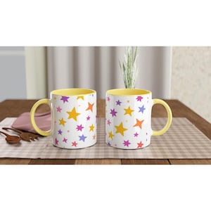 May include: Two white ceramic mugs with yellow interiors and handles. The mugs are decorated with a pattern of colourful stars in orange, yellow, purple, and pink. The mugs are on a checkered surface.