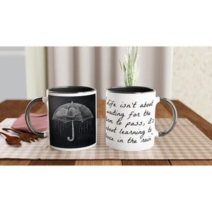 May include: Two white ceramic mugs with black rims. One mug features a black and white illustration of an umbrella with raindrops. The other mug has the text "Life isn't about waiting for the storm to pass, it's about learning to dance in the rain."
