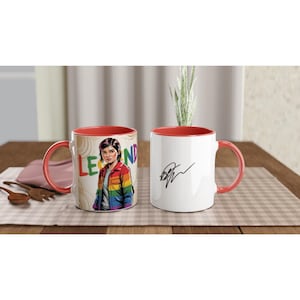 May include: Two white ceramic mugs with red interiors and handles. One mug features a colourful illustration of a person wearing a rainbow jacket, with the word "LEGEND" in rainbow letters. The other mug has a black signature.
