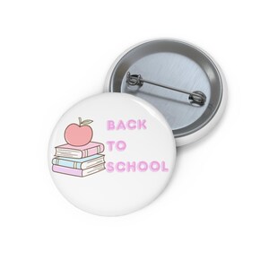 May include: A round white pin-back button featuring a graphic of a pink apple resting on a stack of three books in shades of pink and blue. The text BACK TO SCHOOL is printed in pink capital letters to the right of the books and apple illustration.