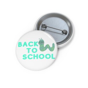 Könnte beinhalten: Ein runder Ansteckbutton mit weißem Hintergrund und dem Text BACK TO SCHOOL in hellgrünen Buchstaben. Neben dem Text befindet sich ein hellgrüner Cartoon-Wurm mit runder Brille. Die Rückseite zeigt einen silbernen Metall-Ansteckmechanismus.