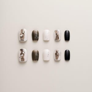 Può includere: Set di dieci unghie finte con una varietà di design. Le unghie sono bianche, nere e marroni con accenti di glitter.