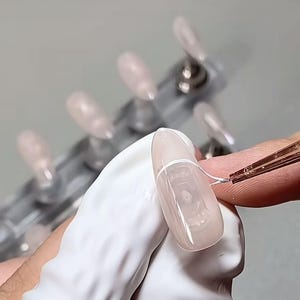 Può includere: Primo piano di una mano con guanto bianco che applica un design a linea bianca su un'unghia acrilica rosa chiaro. L'unghia è di forma ovale. Altre unghie sono visibili sullo sfondo, montate su un supporto trasparente.