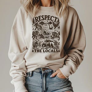 May include: A beige crewneck sweatshirt featuring a dark brown graphic print with the text RESPECT THE LOCALS. The design includes illustrations of cryptids like Bigfoot, Mothman, and the Hodag, along with a map of Wisconsin and forest scenery.