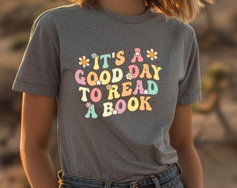 May include: Grey t-shirt featuring the phrase "It's a good day to read a book" in a retro font with floral and star embellishments. The design uses a palette of pastel colours. The t-shirt is made of a soft material.