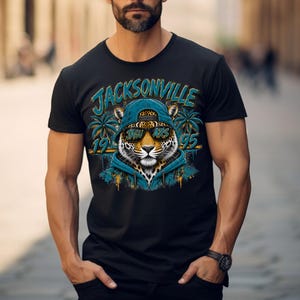 May include: A black short-sleeve t-shirt featuring a graphic of a jaguar wearing a teal hoodie, a backwards baseball cap, and sunglasses. The text reads Jacksonville 1995, with palm trees and paint splatter accents in teal, yellow, and orange.