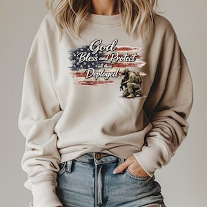 Puede incluir: Sudadera beige con un diseño de la bandera estadounidense y el texto "God Bless and Protect all those Deployed". El diseño incluye una ilustración de un soldado arrodillado. La sudadera es de estilo casual.