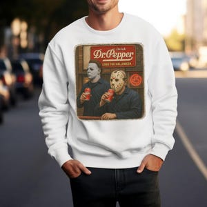 May include: A white crewneck sweatshirt featuring a graphic print of two horror movie characters holding red Dr. Pepper soda cans. Above them, a red sign reads Drink Dr. Pepper GOOD FOR HALLOWEEN. The design includes a neon sign in the background.