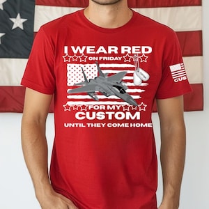 May include: Red t-shirt with a graphic of a fighter jet over an American flag design. The text reads "I Wear Red on Friday for my Custom Until They Come Home". Includes a dog tag graphic.