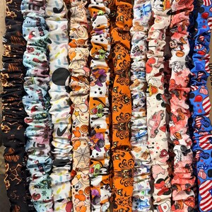 May include: A collection of ten fabric scrunchies arranged vertically, featuring various patterns including black leopard print, Christmas motifs, colorful sprinkles, Halloween spiderwebs, Disney characters, orange pumpkin designs, and patriotic red, white, and blue stars.