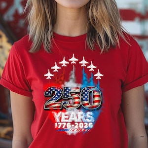 Könnte beinhalten: Rotes T-Shirt mit einem grafischen Design, das 250 Jahre feiert. Das Design enthält die Zahl "250" in einem vom Flaggenstil inspirierten Stil, mit dem Wort "YEARS" und den Daten "1776-2026" darunter. Weiße Jets fliegen über der Zahl.