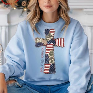 May include: A light blue sweatshirt featuring a patchwork cross design. The cross incorporates American flag patterns, camouflage, and the text "god bless america" in red script. The sweatshirt is a casual top.