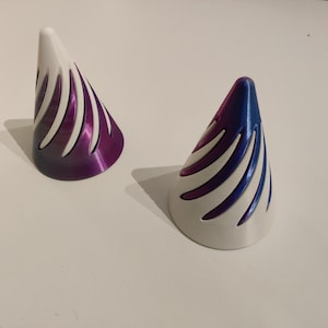 Peut inclure: Deux objets décoratifs en forme de cône. Chacun a une base blanche et un sommet dégradé, l'un violet et l'autre bleu. Les cônes ont des découpes verticales et incurvées. Les objets sont sur une surface blanche.
