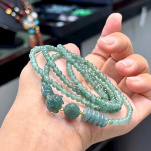 May include: A jade necklace with multiple strands of small, round beads. The necklace features carved jade accents, including a flower, a round bead with a carved design, and a cylindrical bead. The jade has a light green colour.