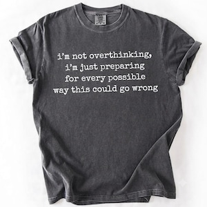 May include: A charcoal grey short-sleeved t-shirt with white text printed on the front that reads: i'm not overthinking, i'm just preparing for every possible way this could go wrong. The shirt features a crew neckline and rolled sleeves.