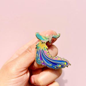 Puede incluir: Un pin de esmalte dorado con un p&aacute;jaro colorido con una cola larga y fluida. El p&aacute;jaro est&aacute; representado en tonos de azul, morado, rosa, amarillo y verde.