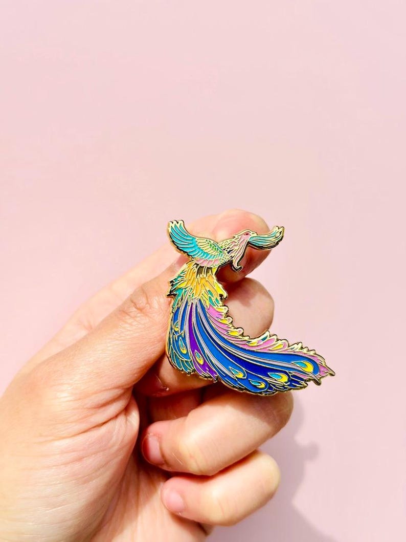 Puede incluir: Un pin de esmalte dorado con un p&aacute;jaro colorido con una cola larga y fluida. El p&aacute;jaro est&aacute; representado en tonos de azul, morado, rosa, amarillo y verde.