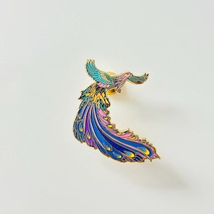 Puede incluir: Un pin de esmalte dorado con un p&aacute;jaro colorido con una cola larga y fluida. El p&aacute;jaro est&aacute; representado en vuelo con las alas extendidas y las plumas de la cola arrastr&aacute;ndose detr&aacute;s de &eacute;l. El cuerpo del p&aacute;jaro es una mezcla de amarillo, rosa y azul, mientras que sus plumas de la cola son un degradado de azul, morado y rosa.