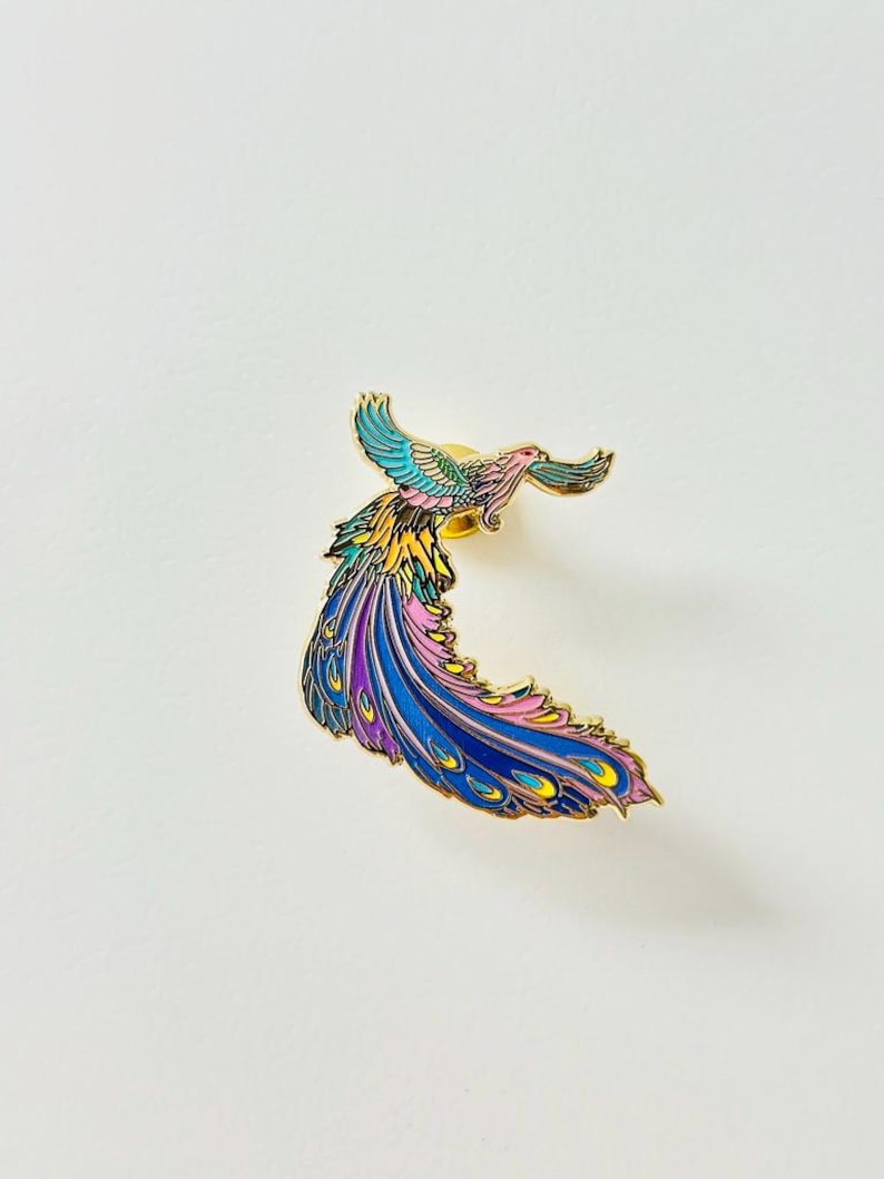 Puede incluir: Un pin de esmalte dorado con un p&aacute;jaro colorido con una cola larga y fluida. El p&aacute;jaro est&aacute; representado en vuelo con las alas extendidas y las plumas de la cola arrastr&aacute;ndose detr&aacute;s de &eacute;l. El cuerpo del p&aacute;jaro es una mezcla de amarillo, rosa y azul, mientras que sus plumas de la cola son un degradado de azul, morado y rosa.